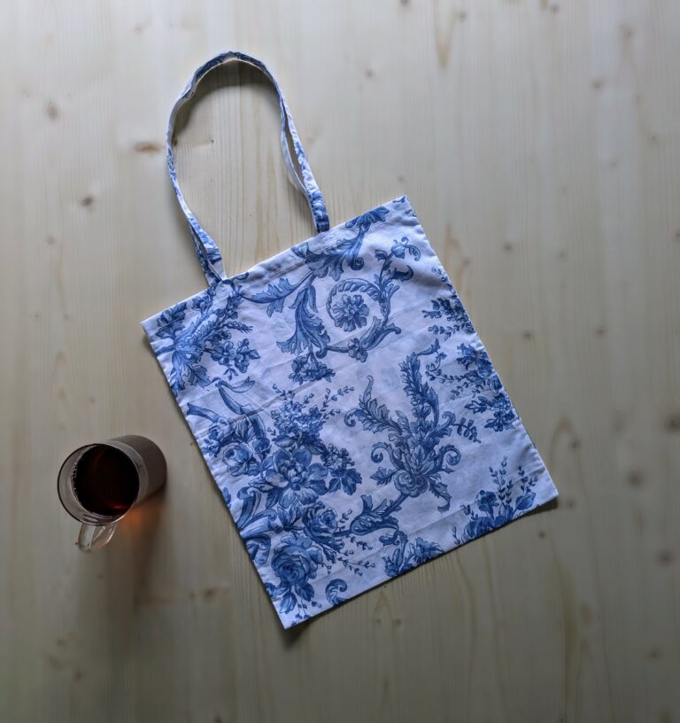 un tote bag en tissu blanc imprimé de fleurs bleues type toile de jouy est posé sur une table avec une tasse de thé à sa gauche