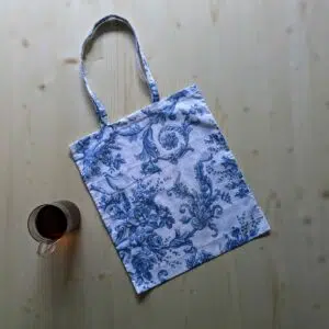 un tote bag en tissu blanc imprimé de fleurs bleues type toile de jouy est posé sur une table avec une tasse de thé à sa gauche