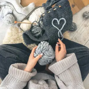 une femme assise tient une pelote de laine grise avec devant elle un tricot gris foncé et un coeur formé avec la laine