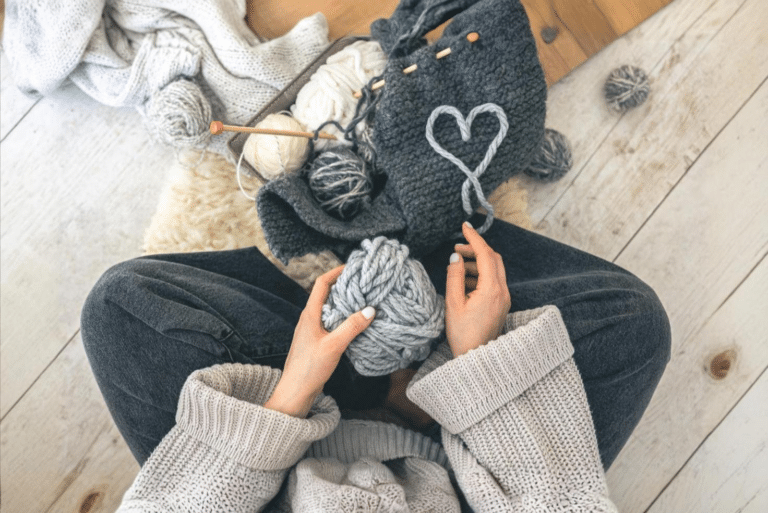 une femme assise tient une pelote de laine grise avec devant elle un tricot gris foncé et un coeur formé avec la laine