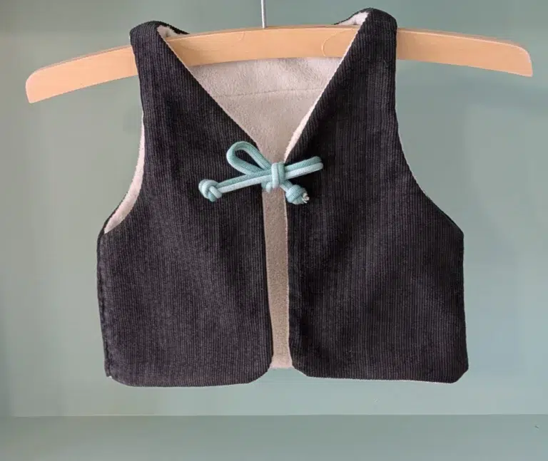 gilet de berger enfant réalisé avec des tissus upcyclés, la face présentée est en velours noir, l'intérieur est en polaire écrue, le lien des deux pans avant est un cordon double avec une partie vert clair et une partie vert plus foncé