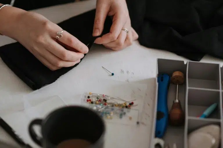 des mains de femme épingle un tissu noir sur la table sont disposées des épingles à tête de verre de couleur une tasse et divers accessoires de couture