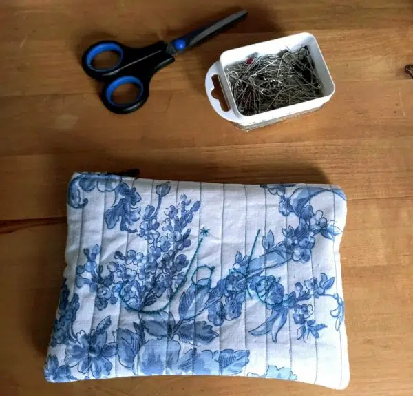 une trousse matelassée en tissu blanc avec des fleurs bleues style toile de jouy posée sur une table en bois clair au dessus de la trousse on voit une boite d'épingles rectangulaire blanche et une paire de ciseaux noir et bleu