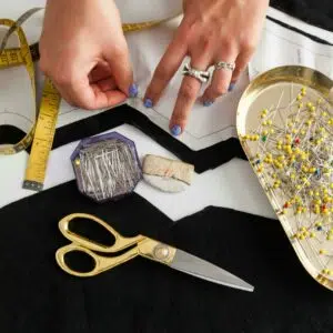 les mains d'une femme dont la main gauche porte 2 bagues épinglent un patron sur un tissu noir. il y a une boite d'épingle à têtes de verre coloré à droite de l'image un mètre ruban jaune à gauche de l'image et au centre, une craie tailleur, des ciseaux dorés et une boite octogonale d'épingles