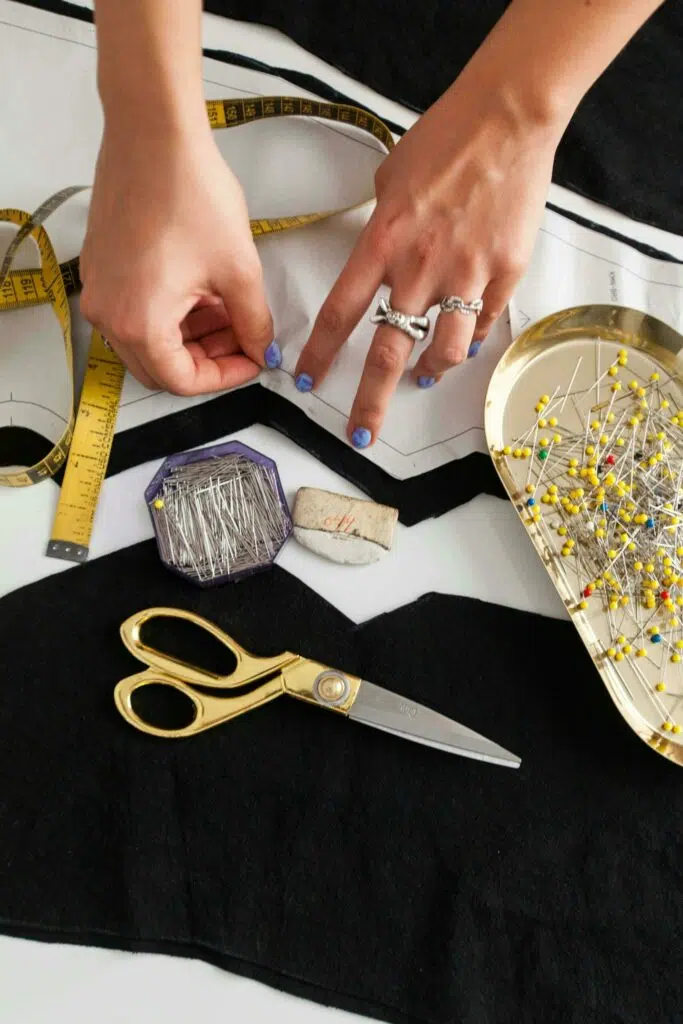 les mains d'une femme dont la main gauche porte 2 bagues épinglent un patron sur un tissu noir. il y a une boite d'épingle à têtes de verre coloré à droite de l'image un mètre ruban jaune à gauche de l'image et au centre, une craie tailleur, des ciseaux dorés et une boite octogonale d'épingles